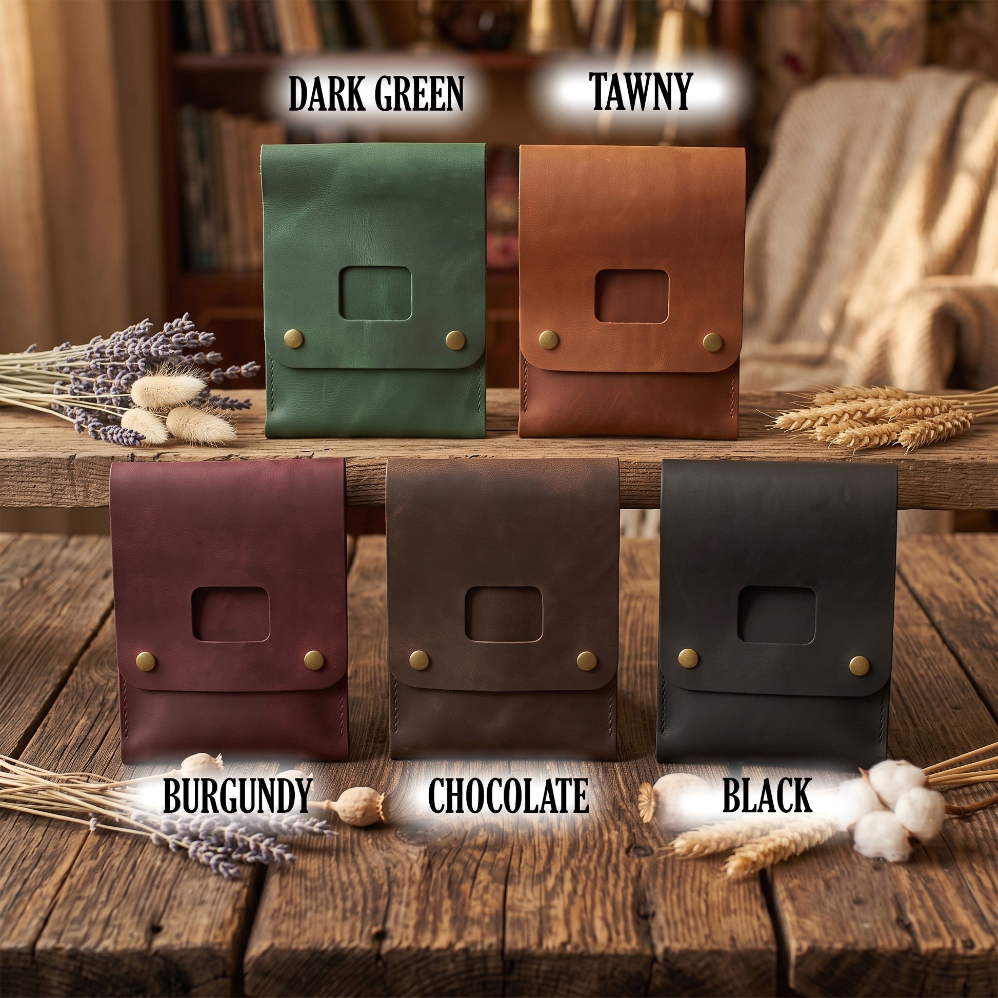 Six leather pouches or small cases shown in different colors on a rustic wooden table: dark green, tawny, burgundy, chocolate, and black. Each has a flap closure with two brass-tone studs and a rectangular cutout.