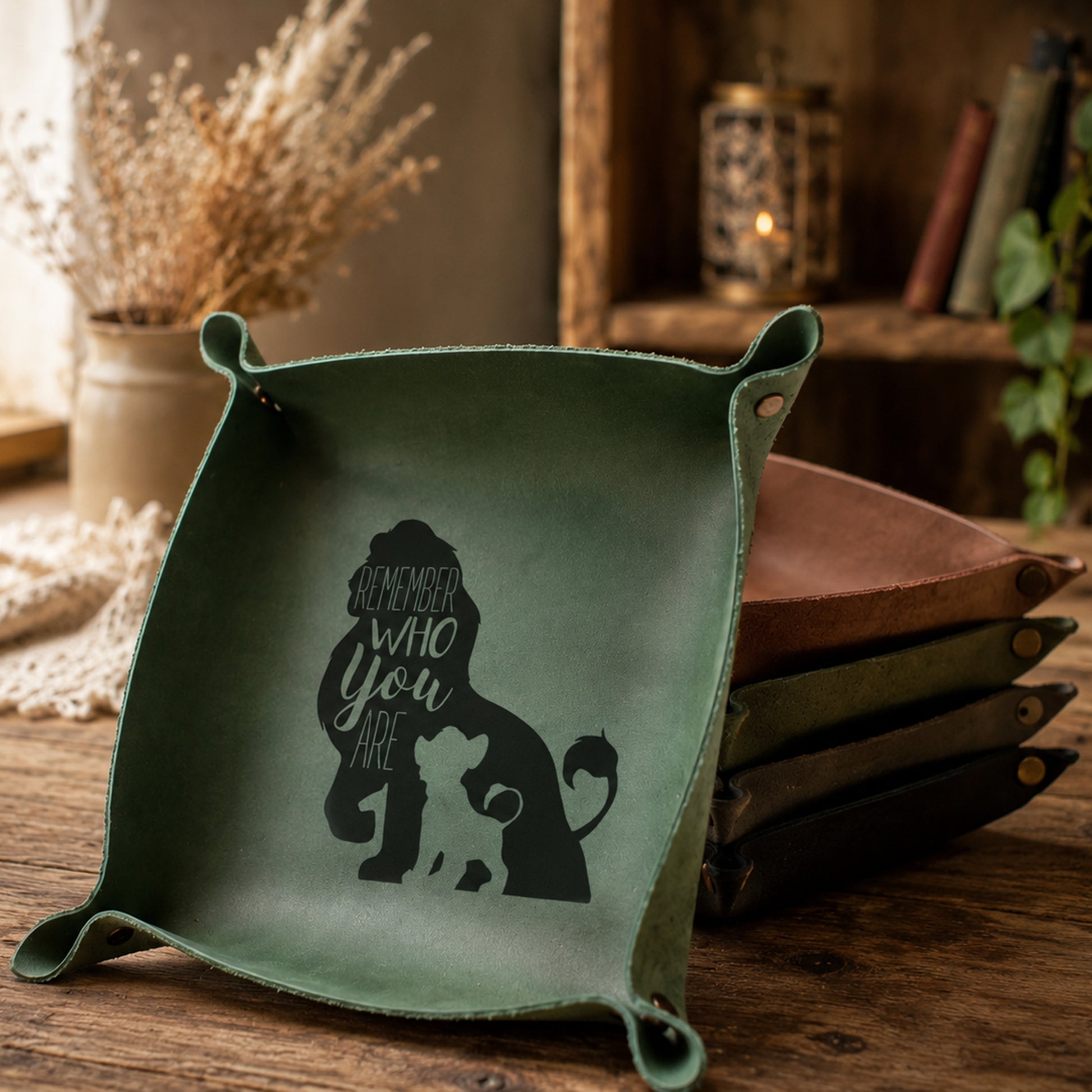 A square green leather dog tray with corner tabs and a black silhouette graphic of a lion and cub with the text 'Remember who you are' inside, displayed on a rustic wooden table with other leather trays in the background.