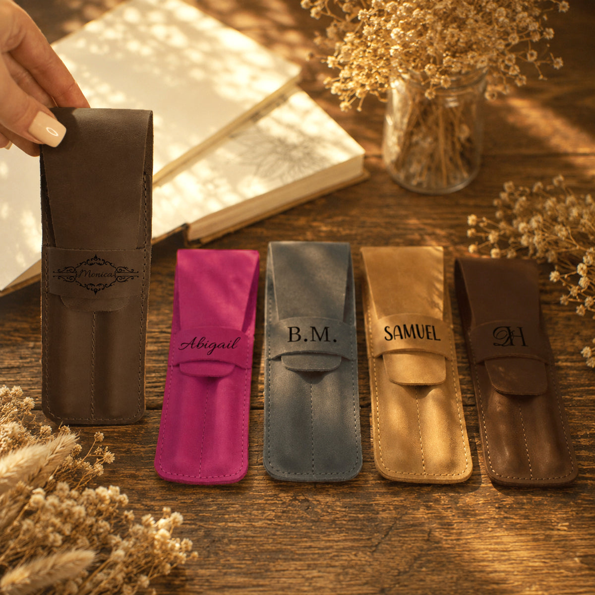 Five personalized leather pen sleeves in different colors (brown, magenta, slate blue, tan, and dark brown) are lined up on a wooden table with a hand holding a brown sleeve on the left. Each sleeve features a unique monogram or name (Abigail, B.
