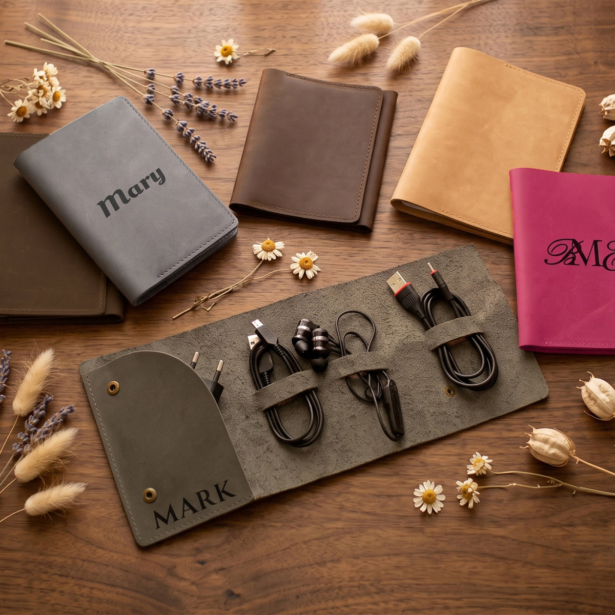 A flat-lay of personalized leather organizer sleeves on a wooden table, styled with dried flowers. The set includes a gray sleeve with 'Mary' and several other sleeves in brown, tan, and magenta, along with matching black USB charging cables.