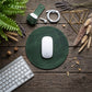 A dark green round mouse pad with a white Apple Magic Mouse on top, styled in a flat-lay on a rustic wooden table with a keyboard, earbuds, and dried plants.