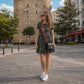 Woman in a floral dress with sunglasses and a black bag walking on a street with a tower in the background.