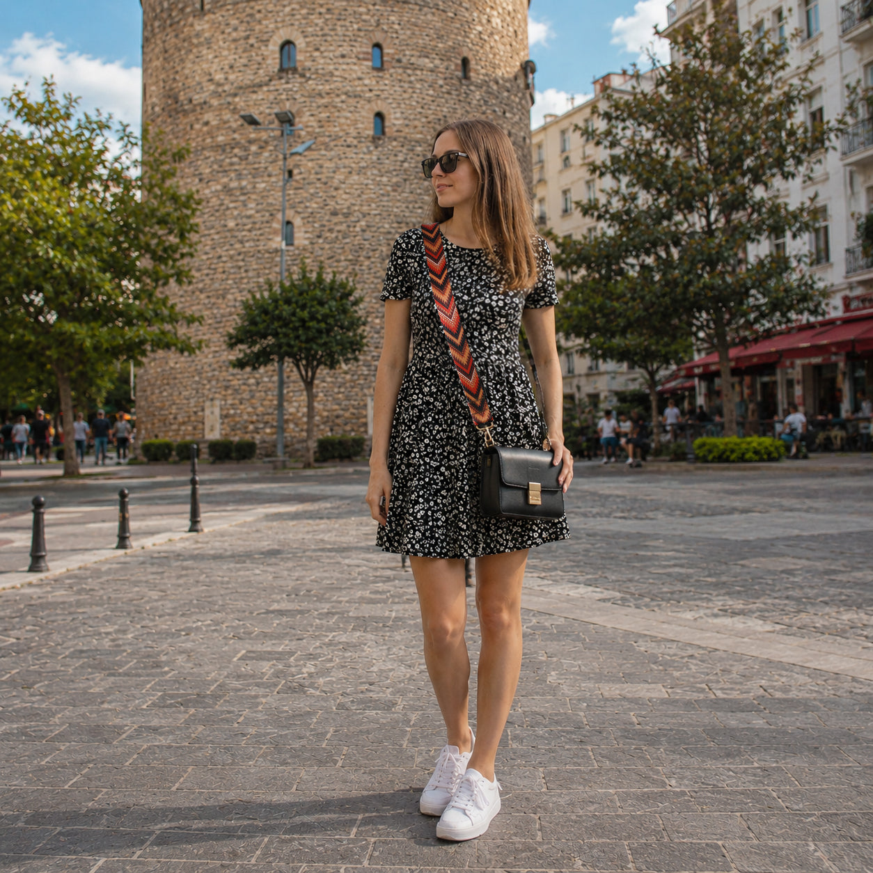 Woman in a floral dress with sunglasses and a black bag walking on a street with a tower in the background.