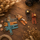 Four moose-shaped keychains in different colors (blue, brown, tan, and dark brown) arranged on a rustic wooden table with dried flowers, coins, and a camera lens prop.