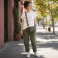 Woman standing on a sidewalk wearing a white t-shirt, green pants, and carrying a gray bag with an orange strap.