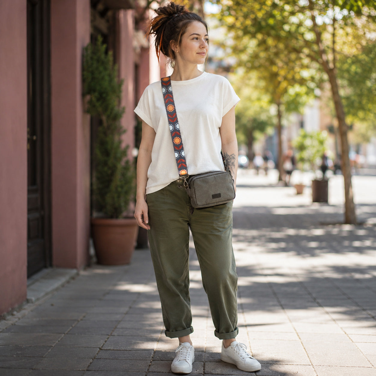 Woman standing on a sidewalk wearing a white t-shirt, green pants, and carrying a gray bag with an orange strap.