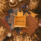A leather tray organizer in a warm mustard-yellow color, personalized with the name 'AMELIA' in black lettering. It is displayed on a rustic wooden table surrounded by other leather trays in brown and black, dried flowers, keys, a watch, and a glass jar with fairy lights.
