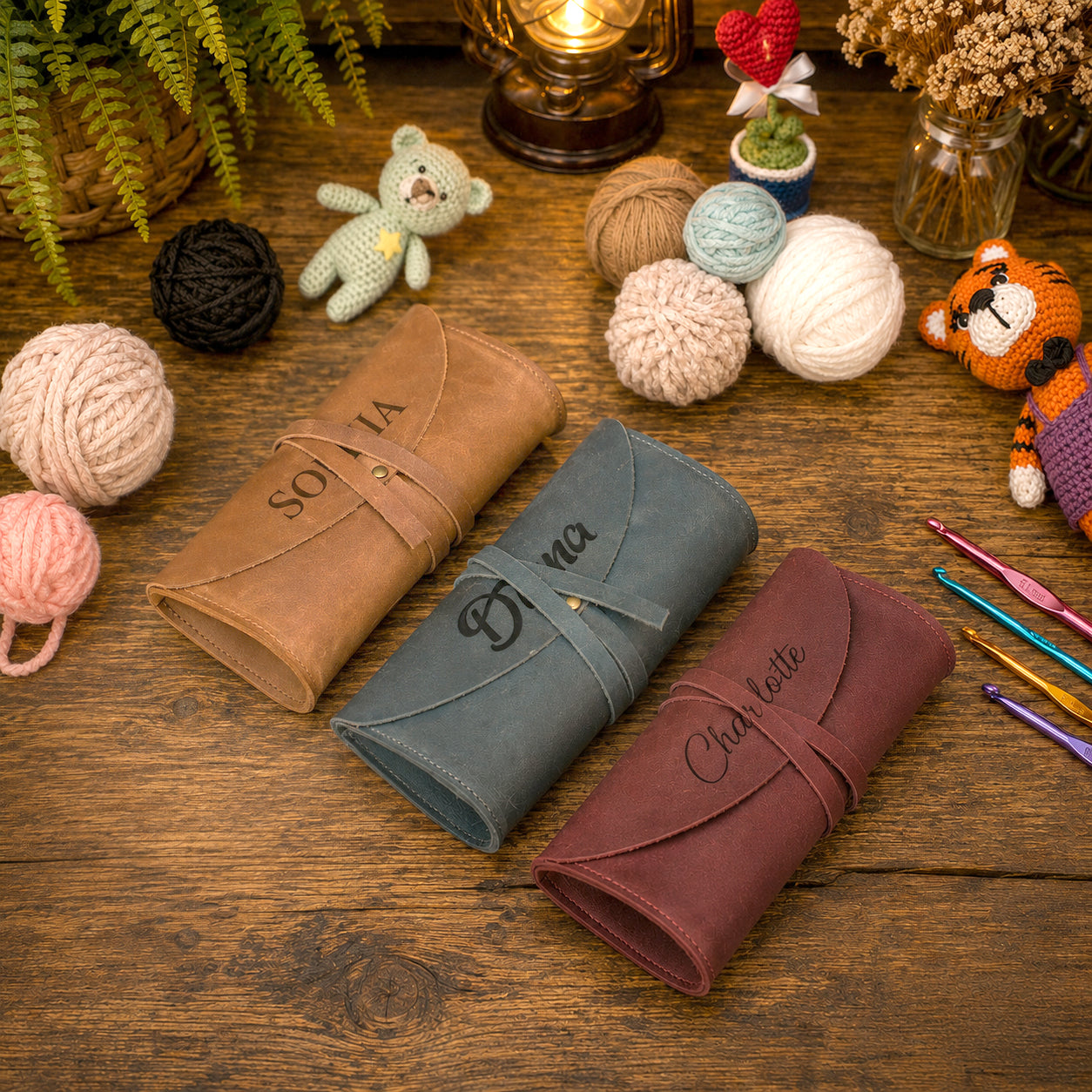 Three rolled leather journal covers in tan, slate blue, and burgundy are arranged on a rustic wooden table, each with a strap closure and a name engraved on the front: Sofia, Bella, and Charlotte. The scene is styled with yarn balls, crochet hooks, a small teddy bear, plants, and warm ambient decor in the background.