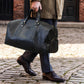 Person holding a black leather duffel bag with 'DS' initials on a cobblestone street.