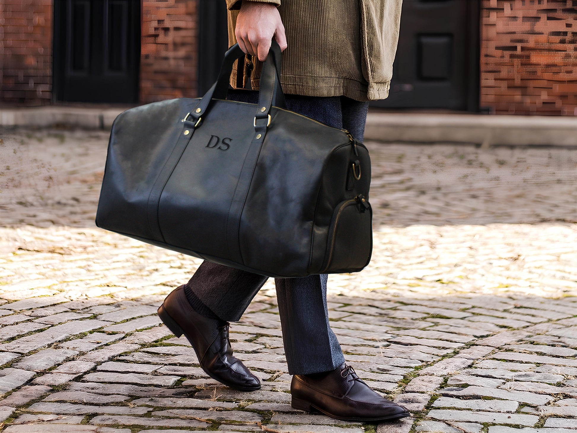 Person holding a black leather duffel bag with 'DS' initials on a cobblestone street.