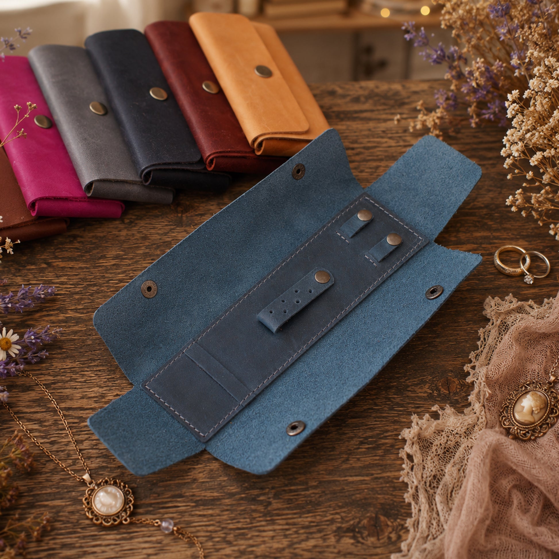 A blue velvet ring roll with brass-tone snaps is displayed open on a rustic wooden table, surrounded by dried flowers, jewelry, and other ring rolls in pink, gray, navy, and tan. No brand is visible.