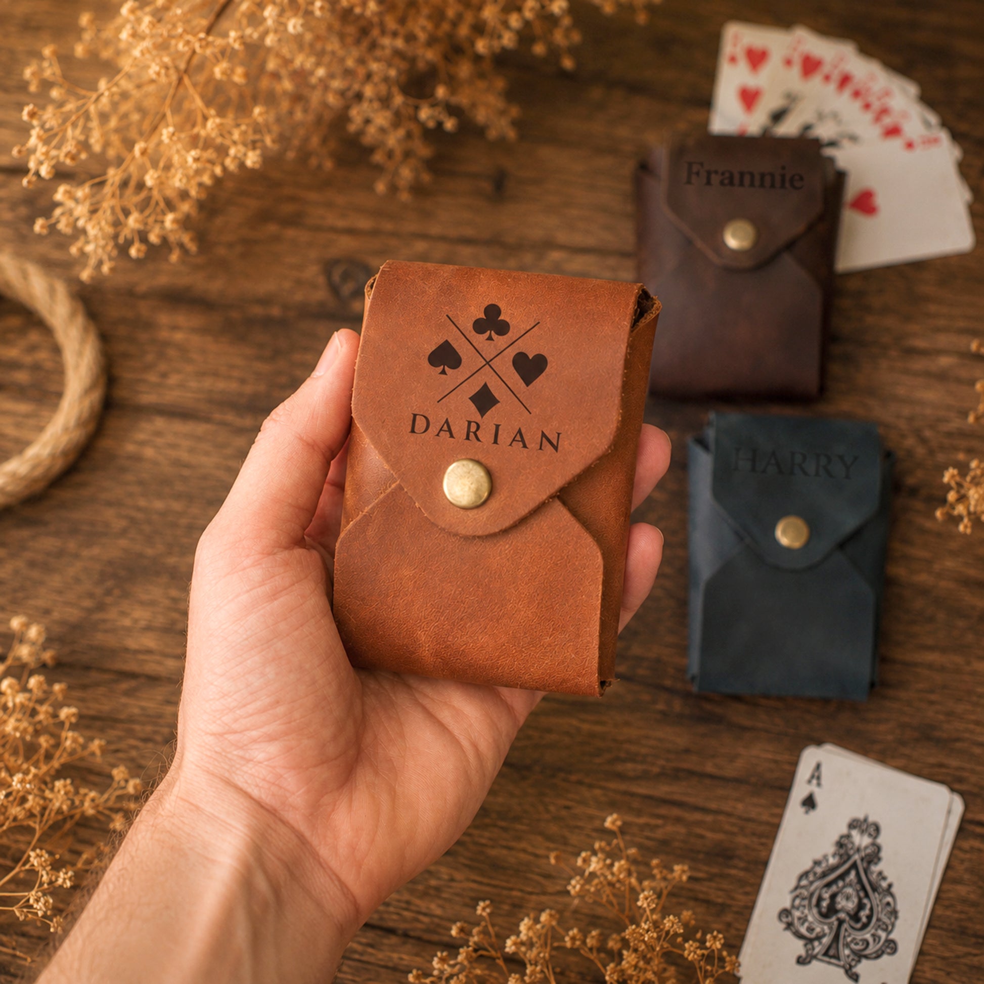A hand holds a small brown leather playing card pouch engraved with a club and diamond symbol and the name DARIAN. In the background on a wooden surface are two more personalized card pouches, playing cards, and dried flowers.