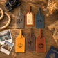 Six personalized leather luggage tags in various colors (gray, beige, blue, yellow, brown, and reddish-brown) are arranged in a flat-lay on a rustic wooden table with travel-themed props including a camera, rope, instant photos, coins, a lantern, and a passport.