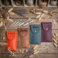 Four personalized leather pouches in orange, tan, teal, and burgundy, each with a snap button and a name engraved on the flap, arranged on a rustic wooden surface with makeup brushes and dried flowers.