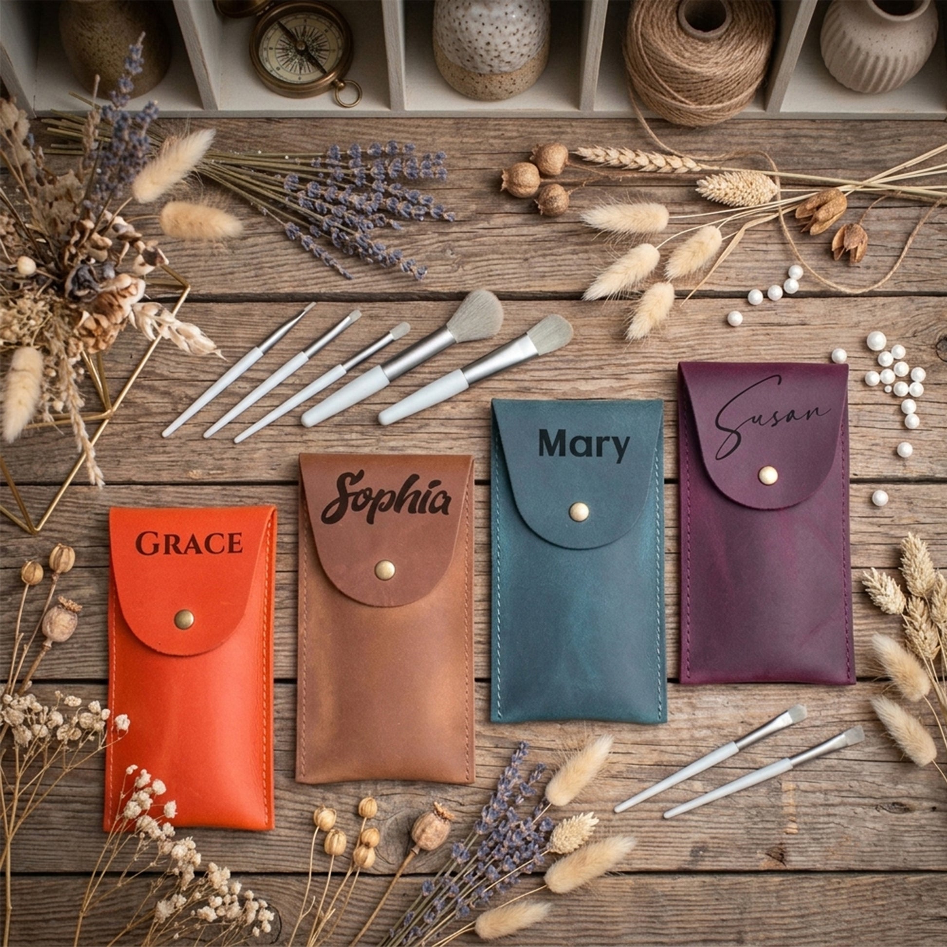 Four personalized leather pouches in orange, tan, teal, and burgundy, each with a snap button and a name engraved on the flap, arranged on a rustic wooden surface with makeup brushes and dried flowers.