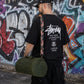 Person wearing a black Stussy t-shirt with a colorful bag against a graffiti-covered wall.