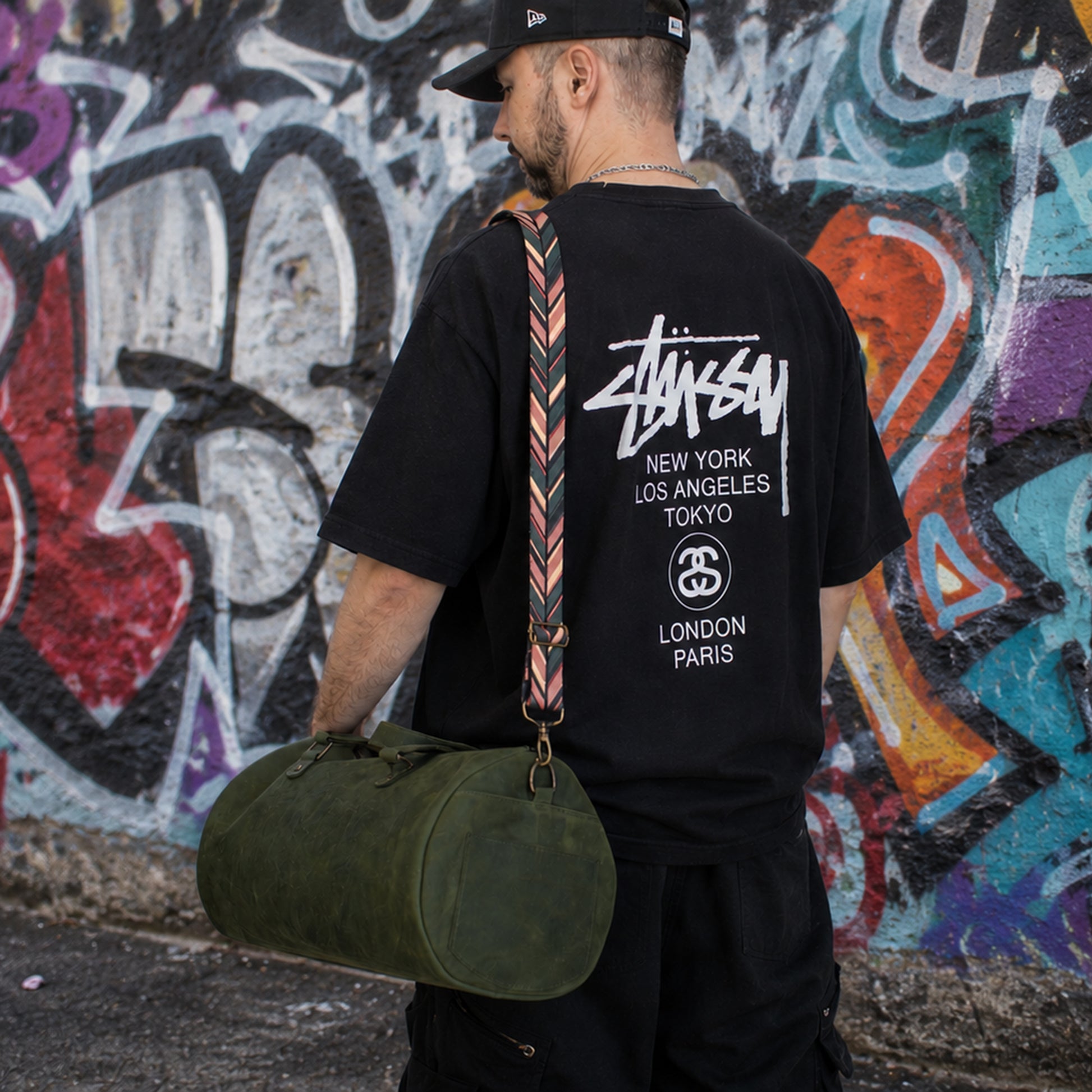 Person wearing a black Stussy t-shirt with a colorful bag against a graffiti-covered wall.