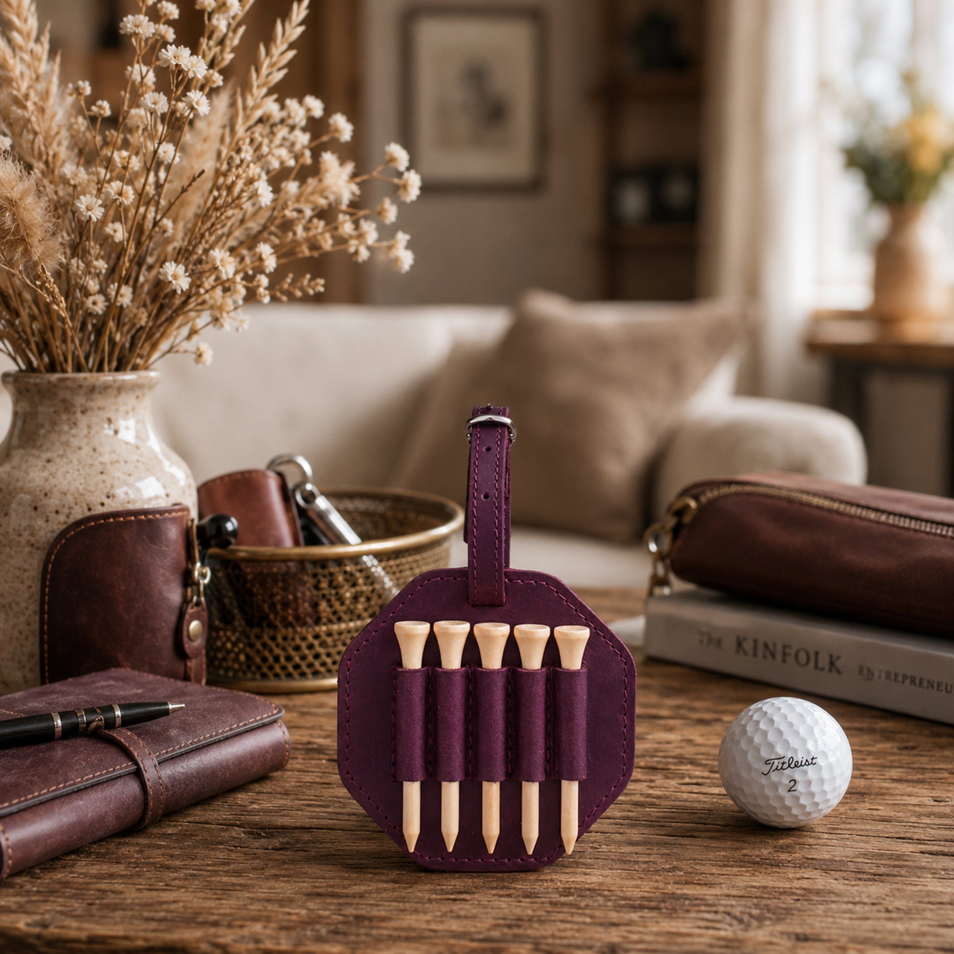 A purple leather golf tees roll holding five wooden golf tees, displayed on a wooden table in a cozy living room setting with a golf ball, books, and leather accessories.