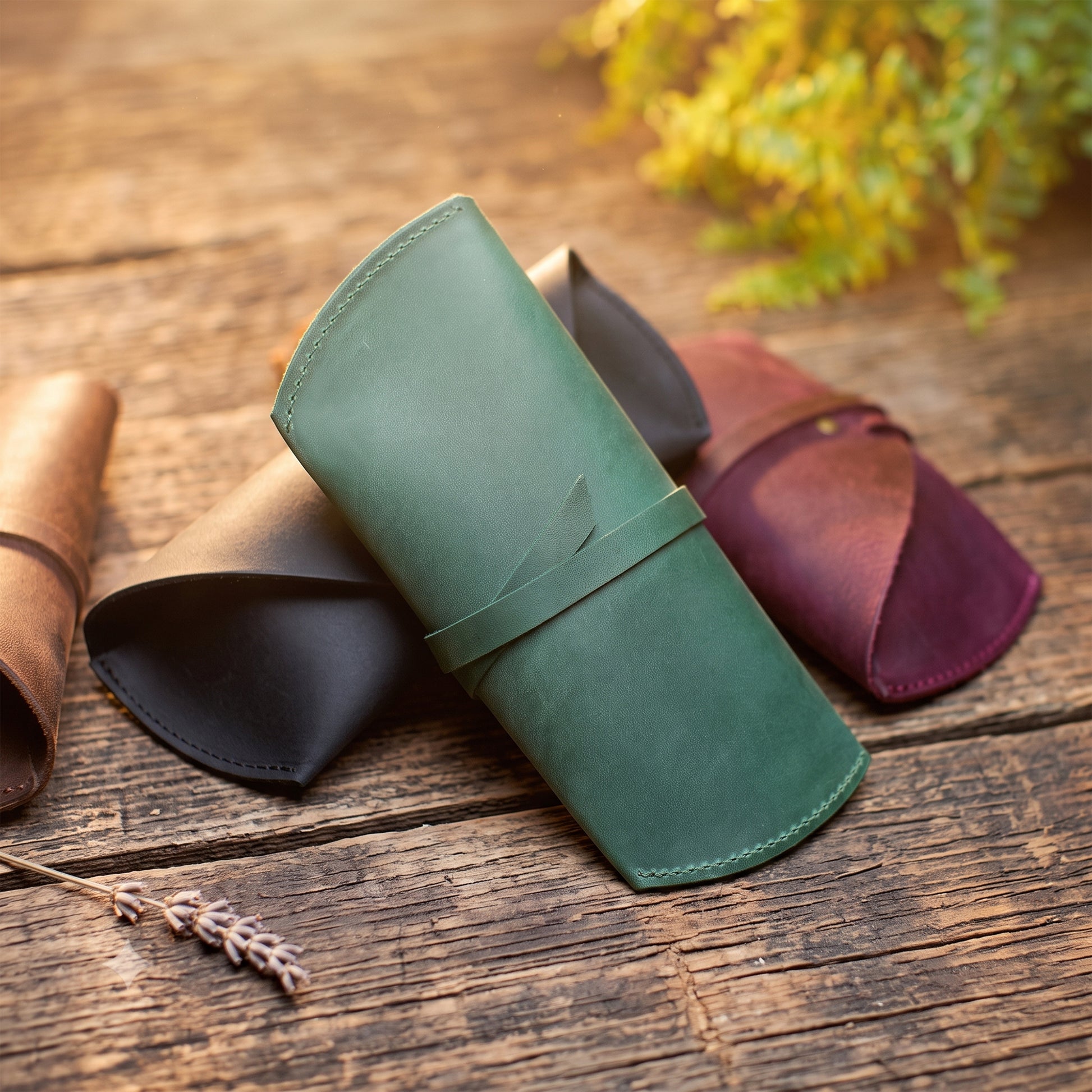 A rolled green leather case or sleeve is displayed on a rustic wooden surface, with two additional rolled leather pieces in brown and burgundy behind it. A sprig of dried lavender and blurred greenery appear in the foreground and background.