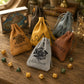 Five personalized drawstring pouches in tan, mustard yellow, dark brown, dusty blue, and light gray velvet, arranged on a rustic wooden table with dice and books around them. The front pouch is light gray and personalized with the name 'Alastair' and a black crest, while the others feature monograms and names like Dominic, Daniel, and Michael.
