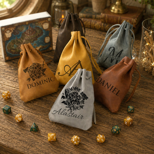 Five personalized drawstring pouches in tan, mustard yellow, dark brown, dusty blue, and light gray velvet, arranged on a rustic wooden table with dice and books around them. The front pouch is light gray and personalized with the name 'Alastair' and a black crest, while the others feature monograms and names like Dominic, Daniel, and Michael.