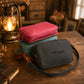 A stack of three personalized canvas zippered pouches sits on a rustic wooden table. The top pouch is a vibrant pink, the middle is teal, and the bottom one is dark gray with the name 'THOMAS' printed on it.