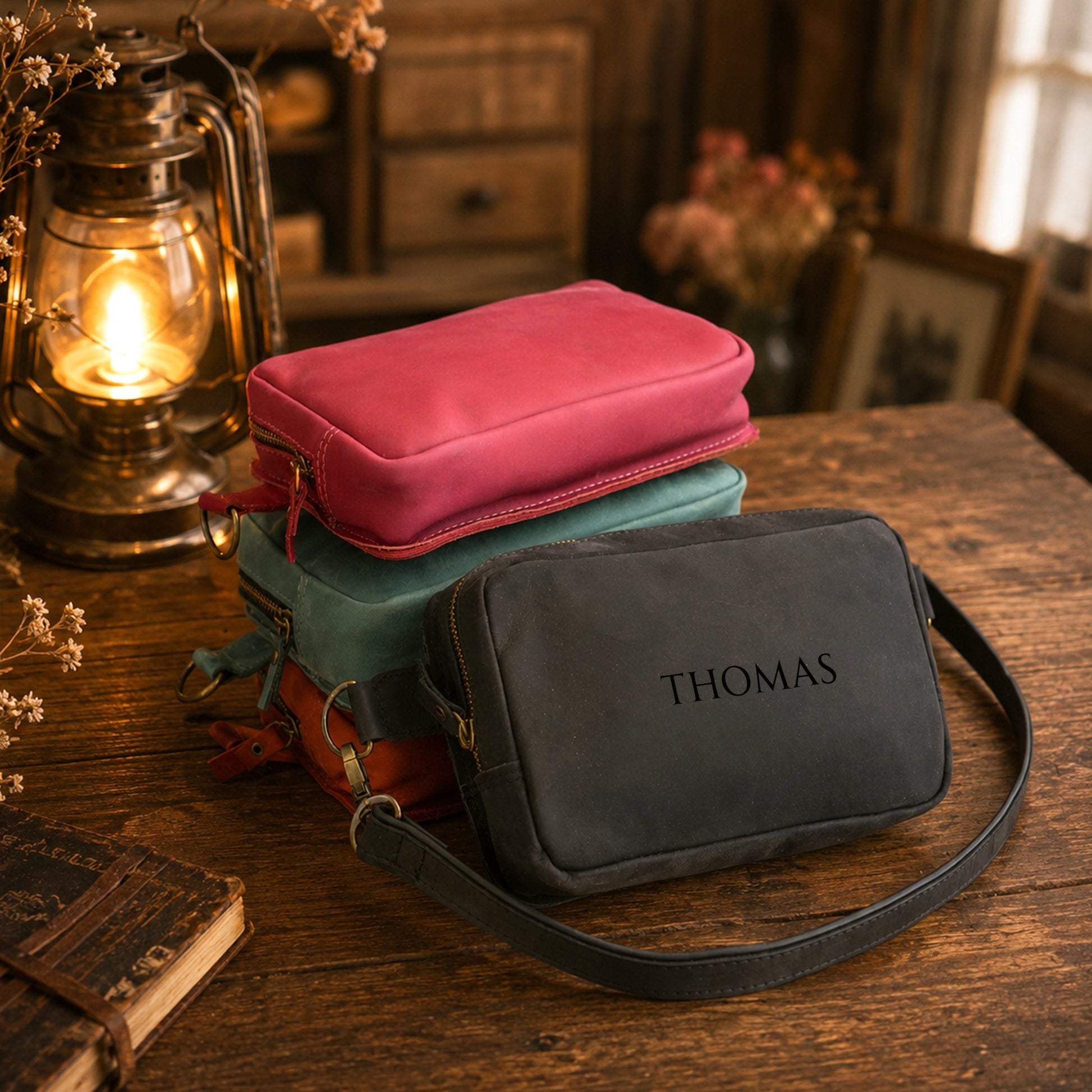 A stack of three personalized canvas zippered pouches sits on a rustic wooden table. The top pouch is a vibrant pink, the middle is teal, and the bottom one is dark gray with the name 'THOMAS' printed on it.