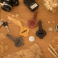 Four personalized guitar-shaped keychains in different colors (yellow, brown, black, and gray) with engraved names or monograms, arranged on a wooden tabletop with vintage props like a camera, keys, coins, and a map.