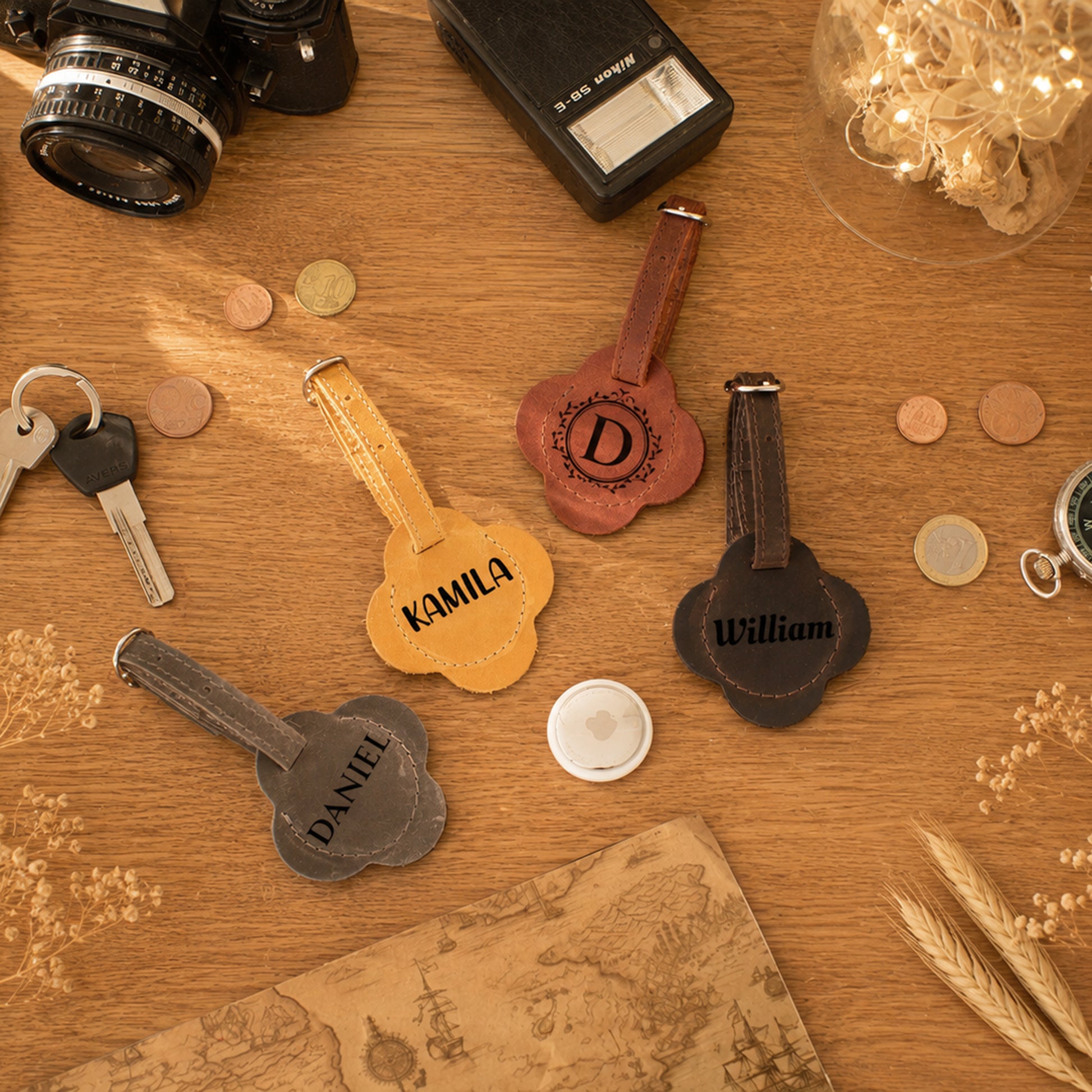 Four personalized guitar-shaped keychains in different colors (yellow, brown, black, and gray) with engraved names or monograms, arranged on a wooden tabletop with vintage props like a camera, keys, coins, and a map.