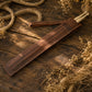 A brown leather strap or sleeve holding two light wooden sticks, arranged diagonally on a rustic dark wood surface with dried flowers and rope as styled props.