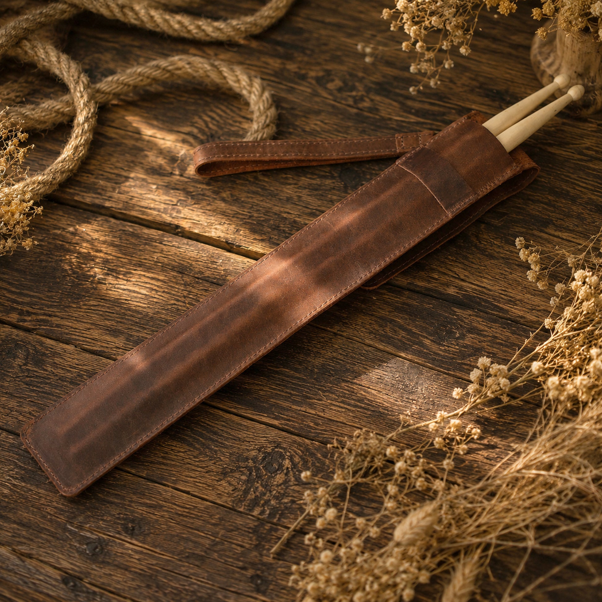 A brown leather strap or sleeve holding two light wooden sticks, arranged diagonally on a rustic dark wood surface with dried flowers and rope as styled props.