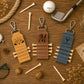 Three personalized leather golf keychains with metal clasp hooks are arranged on a wooden tabletop with golf-themed props including tees, a golf ball, a golf club, rope, and dried flowers. The keychains are tan, brown, and slate blue, each with engraved names or monograms and small wooden tee accents.