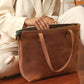 Brown leather tote bag with 'Olivia' branding held by a person in a white outfit.
