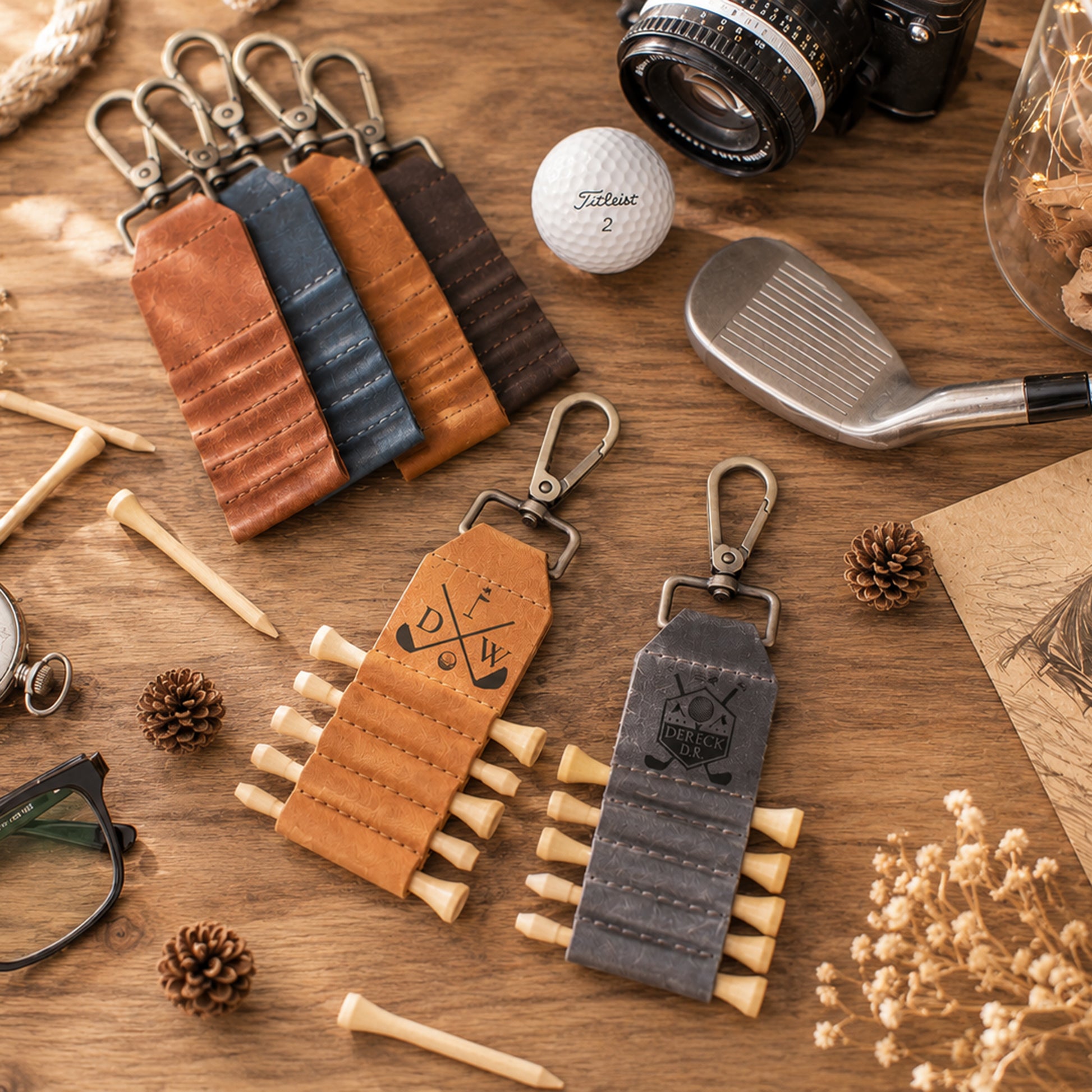 Two leather golf keychains are arranged on a wooden tabletop with golf-themed props, including a golf ball, club, tees, pinecones, and eyeglasses. One keychain is tan and the other is dark gray, both with metal keyrings and a roll of faux tees.