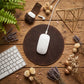 A white Apple Magic Mouse rests on a round dark brown leather mouse pad, arranged in a styled flat-lay on a rustic wooden desk with a keyboard, lavender, dried flowers, and decorative accessories.