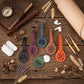 Five personalized leather luggage tags in different colors (black, purple, blue-gray, brown, green, and orange) with monogrammed initials, arranged on a rustic wooden table with travel-themed props like a compass, leather journal, earbuds, and dried lavender.