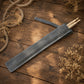 A dark gray leather drumstick sleeve lying diagonally on a rustic wooden surface, with two light wood drumsticks partially visible inside. Dried floral arrangements and rope decorate the corners of the scene.