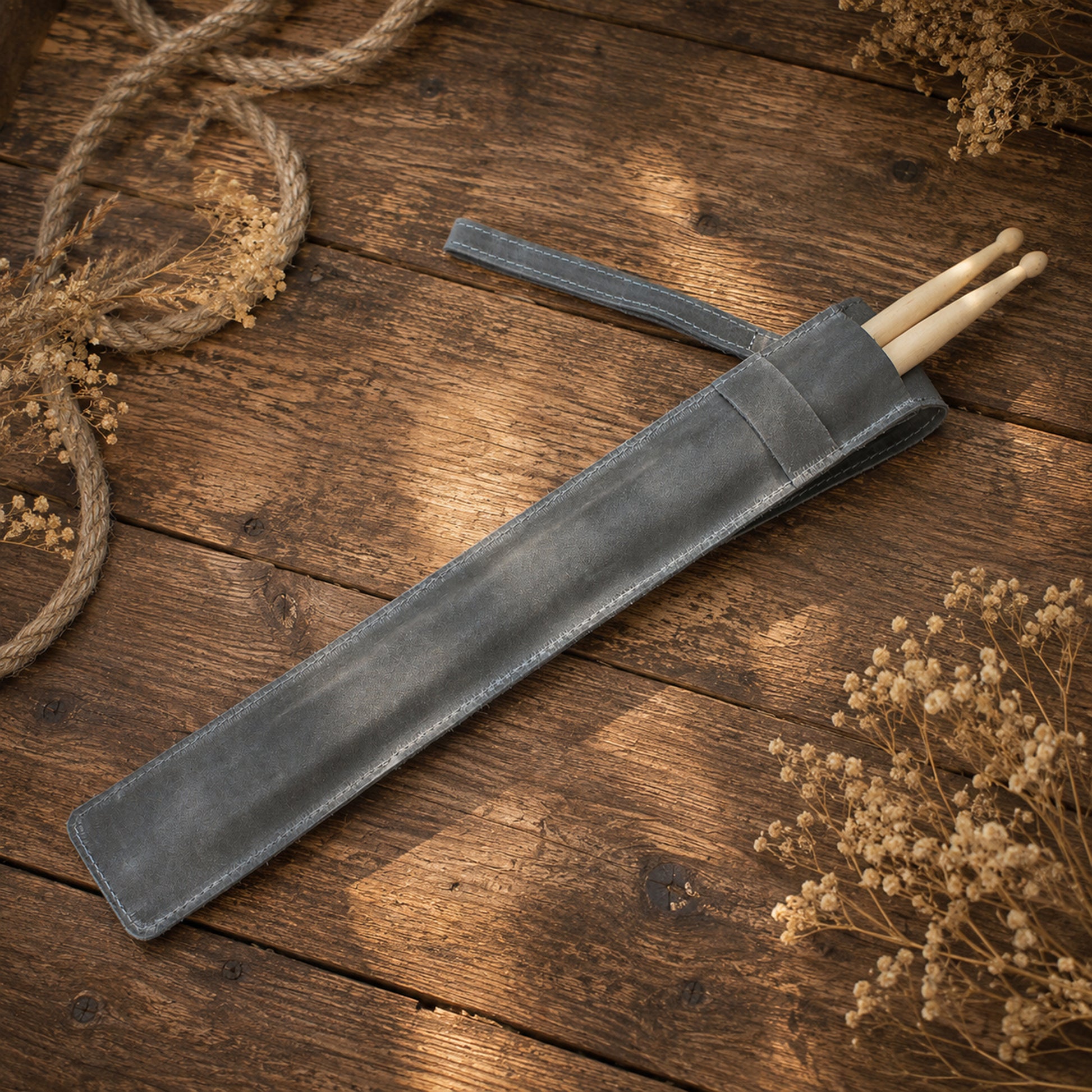 A dark gray leather drumstick sleeve lying diagonally on a rustic wooden surface, with two light wood drumsticks partially visible inside. Dried floral arrangements and rope decorate the corners of the scene.