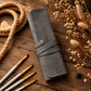 A gray roll-up canvas or fabric storage case with a snap strap and button, styled on a rustic wooden surface with paintbrushes, rope, and dried flowers.