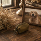 A small olive green canvas zipper pouch with a brass key ring, styled on a rustic wooden table next to a framed photo of a golden retriever, with dried flowers and books in the background.