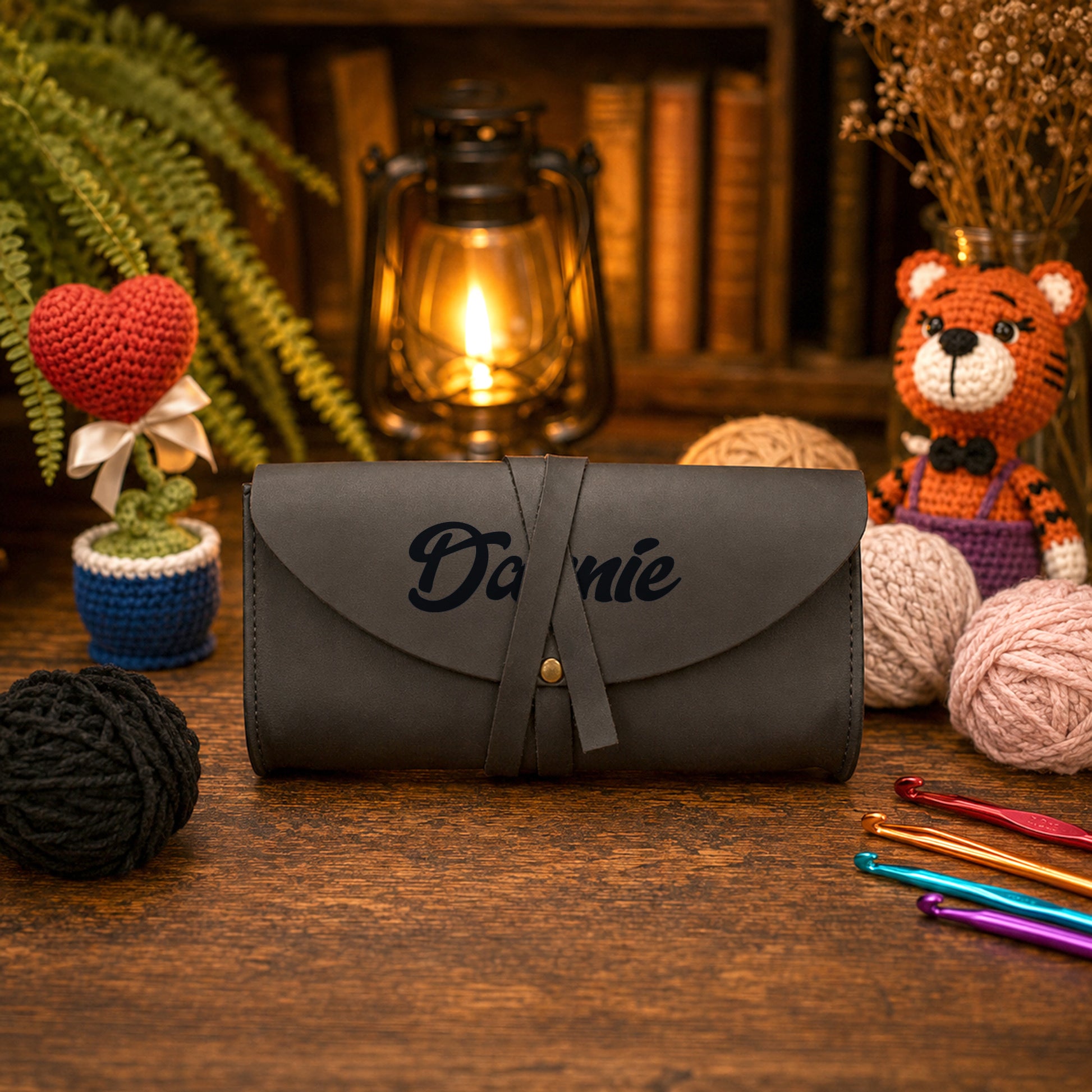 A dark gray personalized crocheting or sewing tool case with the name 'Dannie' embroidered on the front, displayed on a wooden table surrounded by crochet hooks, yarn balls, a crocheted heart plant, and a crocheted tiger doll.
