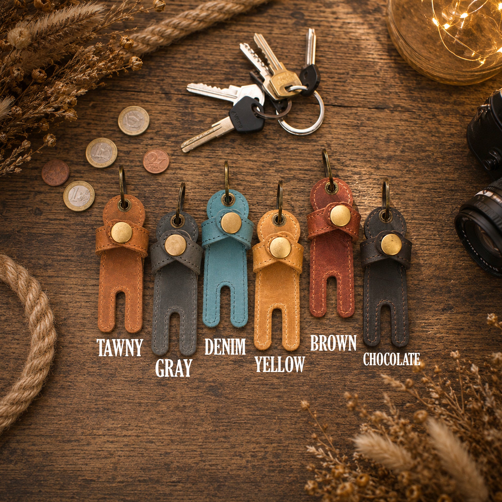 Six leather keychain wallet charms in different colors (Tawny, Gray, Denim, Yellow, Brown, Chocolate) arranged on a rustic wooden table with decorative props like keys, coins, rope, and fairy lights.