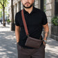 Man wearing a black shirt and brown pants with a brown leather bag on a city street.