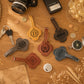 A styled flat-lay of six personalized leather luggage tags on a wooden tabletop, surrounded by vintage props like a camera, keys, coins, a compass, and dried flowers. The tags feature monogrammed initials and names: D, William, Jazzy, A&N, Ivy, and J&K.
