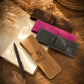 Four personalized leather pen sleeves in brown, magenta, dark gray, and tan, arranged on a wooden desk next to an open notebook and a black pen.