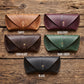 Five leather pouches or cases arranged in a 2x3 grid on a rustic wooden surface, labeled Tawny, Burgundy, Chocolate, Dark Green, and Black. Each pouch has a flap closure with a small metal snap button.