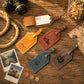Five personalized leather luggage tags in tan, brown, blue, and gray are arranged on a rustic wooden table with vintage props including a camera, rope, coins, and instant photos.