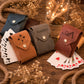 Four personalized card pouches in blue, brown, gray, and reddish-brown leather, each with a snap button and engraved name or monogram, arranged on a wooden surface with playing cards and dried floral decor.
