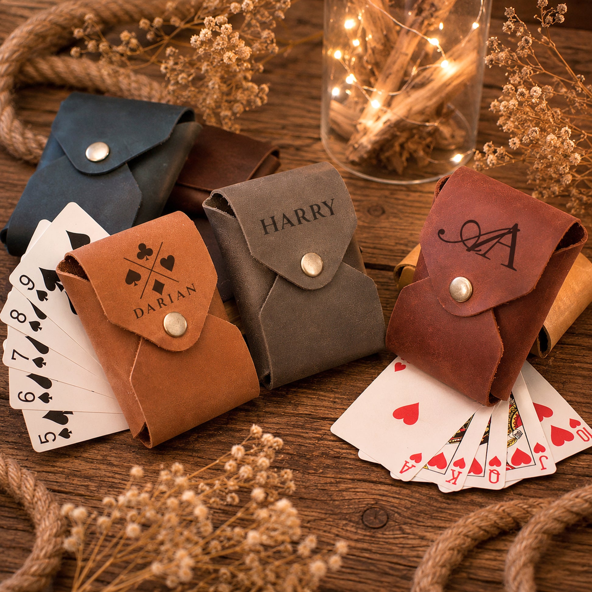 Four personalized card pouches in blue, brown, gray, and reddish-brown leather, each with a snap button and engraved name or monogram, arranged on a wooden surface with playing cards and dried floral decor.