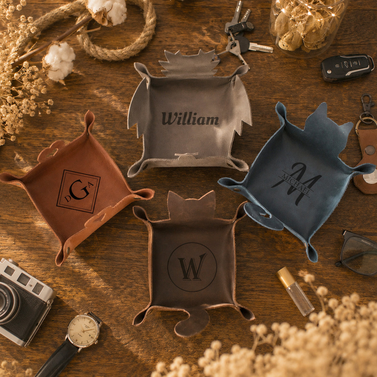 Four personalized leather tray coasters in brown, gray, blue, and bronze, each shaped like a castle with a monogram and name (G, William, M, W) engraved on them, arranged on a wooden table with vintage props.